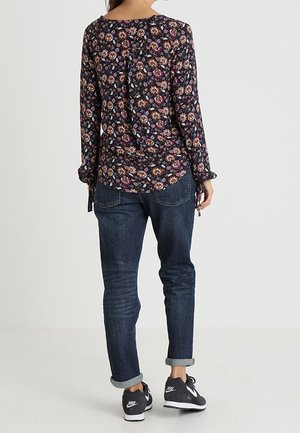 Top floreale a maniche lunghe con sfondo blu navy, cravatte laterali e orlo arrotondato; abbinato a jeans scuri con risvolto e sneakers nere.