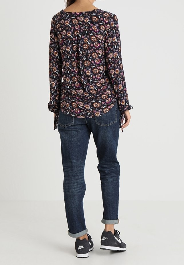Top floreale a maniche lunghe con sfondo blu navy, cravatte laterali e orlo arrotondato; abbinato a jeans scuri con risvolto e sneakers nere.
