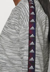 Felpa grigia a righe con un pannello laterale nero che presenta ripetuti loghi Adidas bianchi in un motivo geometrico rosso e blu.