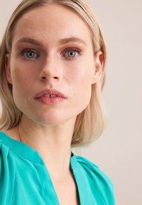 Blusa turchese con spalle plissettate, texture liscia. La pelle della modella mostra un trucco naturale; i capelli chiari sono parzialmente incorniciati. Sfondo neutro.
