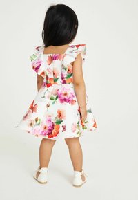 Blumenkleid mit Rüschenärmel, mehrfarbige Blumenmuster auf weißem Hintergrund, figurbetontes Oberteil, schwingender Rock, kombiniert mit weißen Schuhen.
