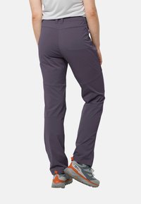 Lila Wanderhosen mit glatter Textur, verstellbarem Bund und vorgeformten Knien, kombiniert mit grauen und orangefarbenen Wanderschuhen.
