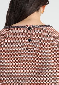 Dzianinowy top z teksturowanym wzorem w kolorach pomarańczowym, białym i czarnym. Posiada okrągły dekolt i trzy ciemne guziki z tyłu.
