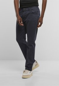 Mann trägt marineblaue, gerade geschnittene Hosen, weiße Turnschuhe und ein schwarzes ärmelloses Oberteil, steht vor einem schlichten grauen Hintergrund.