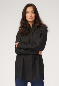 Sort skjorte med knaplukning, krave, lange ærmer og en afslappet pasform. Stoffet ser glat ud med en gloss overflade og subtil glans.