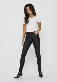 Pikad tumedad juuksed omava naine, kes kannab valget lühikeste varrukatega toppi, musti nahkpükse ja musti kõrge kontsaga ankle saapaid, seisab tasase tausta vastu.