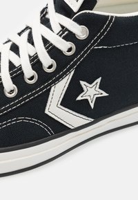 Sneaker alta in tela nera con lacci bianchi, dettaglio a stella e a chevron. Presenta cuciture bianche e una suola in gomma con bordi neri.