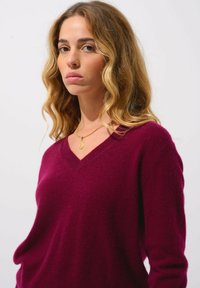 Burgundy v-ringad tröja i mjukt stickat material, med långa ärmar och avslappnad passform. Ett guldhalsband med en berlock är synligt.