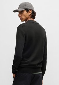 Suéter negro de punto con cuello redondo, puños y dobladillo acanalados. Se lleva una gorra gris, con tejido texturizado y diseño sencillo.