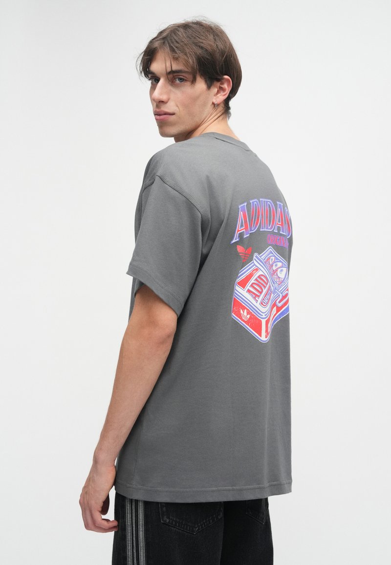 Camiseta de algodón gris de manga corta con estampado gráfico colorido en la parte posterior que incluye el texto "ADIDAS ORIGINAL" y una ilustración de una cinta de casete.