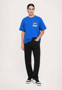 Blå bomull t-skjorte med korte ermer, med et grafisk trykk på brystet. Kombinert med svarte jeans og kraftige svart-hvite joggesko.