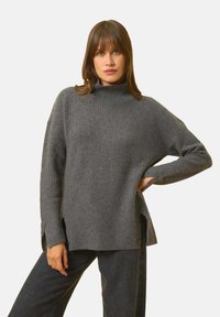 Grauer Rollkragenpullover mit gerippter Textur, seitlichen Schlitzen und weiten Ärmeln, kombiniert mit dunklen Hosen vor einem schlichten weißen Hintergrund.