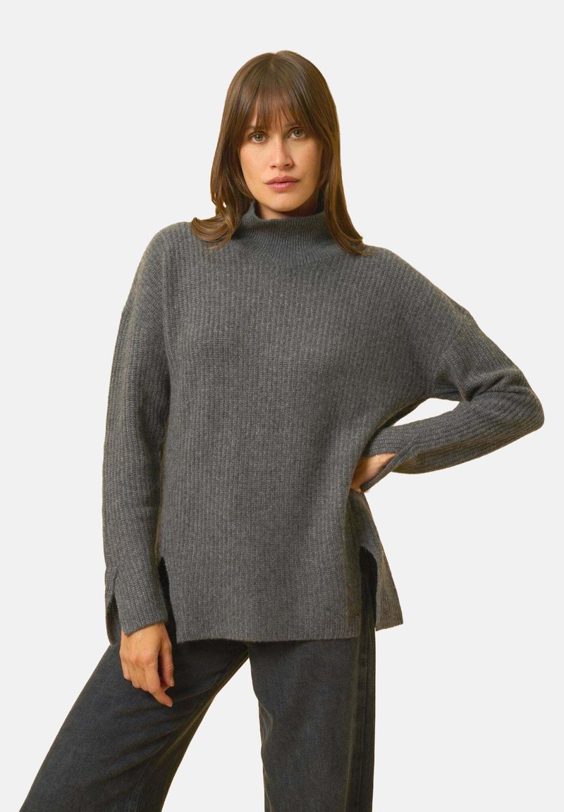 Grauer Rollkragenpullover mit gerippter Textur, seitlichen Schlitzen und weiten Ärmeln, kombiniert mit dunklen Hosen vor einem schlichten weißen Hintergrund.