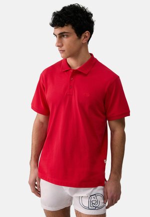 Giovane uomo che indossa una polo a maniche corte rosso brillante e pantaloncini bianchi con una grafica nera su una gamba, in piedi davanti a uno sfondo semplice.
