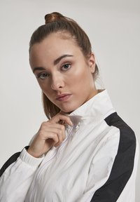 Bijela jakna s visokim ovratnikom i crnim akcentima na ramenima. Ima prednji patentni zatvarač i nabrane manžete s laganom teksturom.