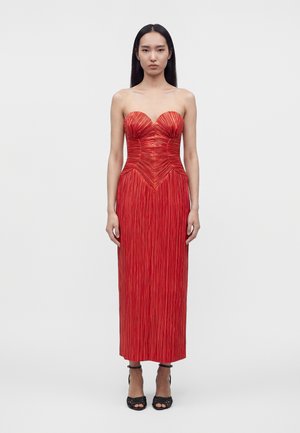 Vrouw staand, gekleed in een strapless, geplooide rode midi-jurk met een sweetheart halslijn en zwarte open-teen hakken tegen een effen achtergrond.