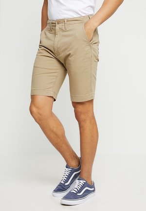 Mand iført beige knælange shorts og mørkeblå sneakers med hvide snørebånd, stående med hænderne i lommerne på en ensfarvet baggrund.