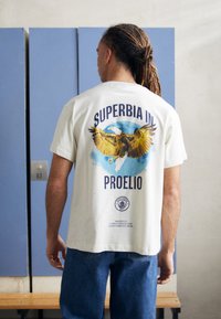 Baltos medvilnės T marškiniai su erelio grafika ir užrašu "SUPERBIA IN PROELIO" tamsiai mėlyna spalva. Trumpomis rankovėmis ir laisvu siluetu.