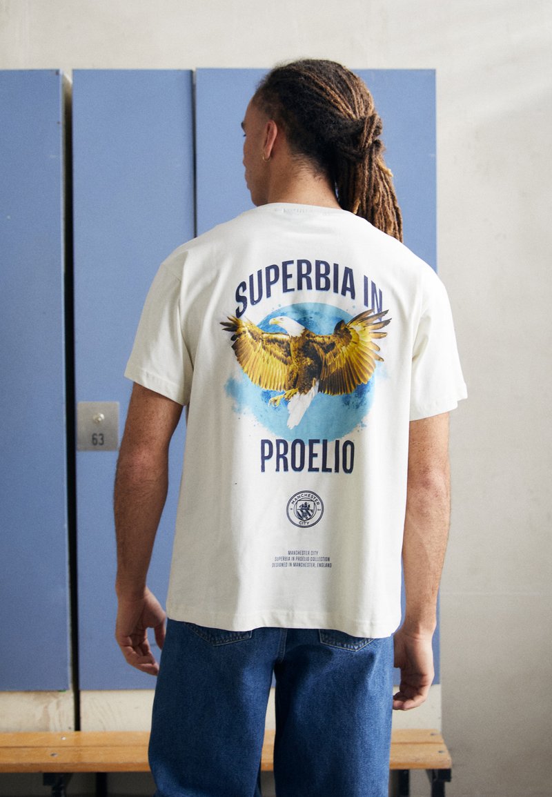 Baltos medvilnės T marškiniai su erelio grafika ir užrašu "SUPERBIA IN PROELIO" tamsiai mėlyna spalva. Trumpomis rankovėmis ir laisvu siluetu.