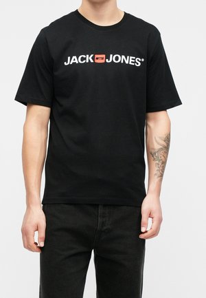 Homme portant un t-shirt noir Jack Jones avec un logo blanc et un carré rouge, associé à un pantalon noir et un tatouage visible sur l'avant-bras.