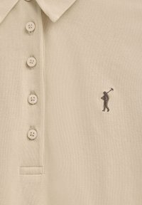 Polo beige con tejido texturizado, tres botones y una pequeña figura bordada en marrón de un golfista en el pecho izquierdo.