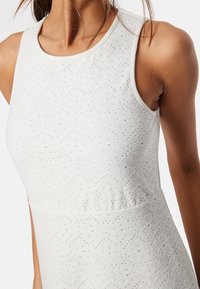 Bílé bezrukávové šaty s texturovaným, vyšitým vzorem a květinovými motivy. Látka se jeví jako lehká a mírně elastická.