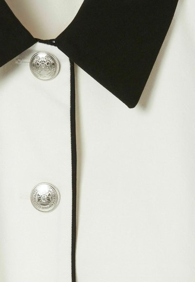 Camisa blanca con cuello negro, que presenta herrajes de botón plateado y ribetes en negro contrastantes a lo largo de la parte delantera. Textura de tela suave.