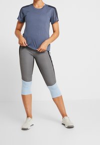 Světle modré sportovní tričko s prodyšnými detaily, spárované se šedými 3/4 legínami s lehce modrými akcenty a černým bočním pruhem.