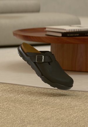 Čierny nazúvací clog so striebornou prackou, položený na béžovom textúrovanom koberci v obývacej izbe s okrúhlym dreveným stolom a knihami.