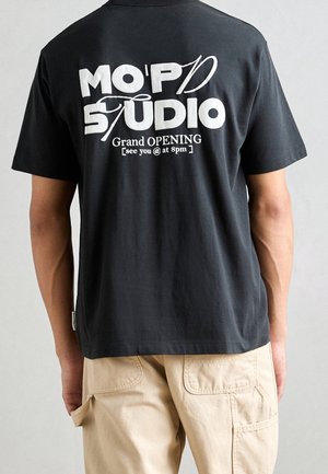 Personne portant un t-shirt noir avec le texte blanc "MO'P STUDIO Grand OPENING [rendez-vous à 20h]" et un pantalon beige, vue de dos.