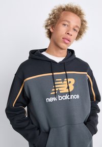 Sudadera con capucha en gris oscuro con mangas negras y un acento dorado. Presenta un gran logo de "New Balance" y un bolsillo frontal. Textura suave.