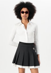 White long-sleeve shirt with a logo on the collar, paired with a black pleated skirt featuring a white hem and a decorative pin. Red sunglasses.