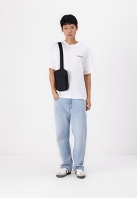 Bolso cruzado negro con un bolsillo frontal con cremallera, llevado sobre una camiseta blanca y unos vaqueros azules. La modelo está de pie sobre un fondo liso.
