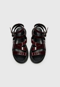 Sandales noires avec des brides en cuir verni bordeaux foncé brillant, présentant un design entrecroisé et une boucle réglable à la cheville. Semelle texturée noire.