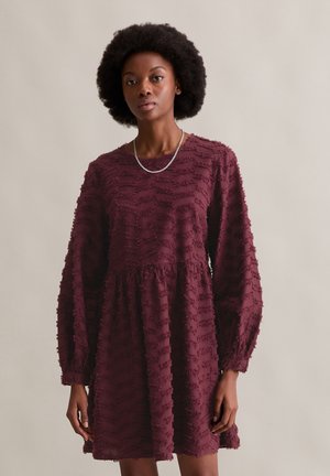 Weinrotes Kleid aus strukturiertem Stoff mit gerafften horizontalen Linien, langen Ärmeln und tailliertem Bund. Schlichter Rundhalsausschnitt und minimalistisches Design.