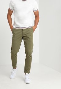 Uomo che indossa una maglietta bianca a maniche corte a coste, pantaloni aderenti verde oliva e sneakers bianche, in piedi con le mani in tasca.