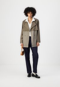 Dobbeltradet, to-farget kåpe i oliven og beige, med store slag og gullknapper. Kombinert med marineblå bukser og svarte mokasiner.