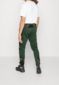 Vihreät verkkarit, joissa on musta ja keltainen sidontavyö, kapeat lahkeet ja sivupaneelin korostukset; yhdistetty valkoiseen T-paitaan ja mustiin lenkkareihin.