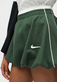 Πράσινη υφασμάτινη φούστα Nike με λευκές λωρίδες και λογότυπο, φορεμένη με λευκό και μαύρο μακρυμάνικο μπλουζάκι, δείχνοντας το μέσο μηρό και το χέρι.