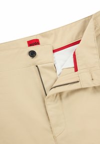 Beige trousers with a black button, red interior lining, and zippered fly. Features a branded tag "HUGO" on the waistband. Durable fabric.