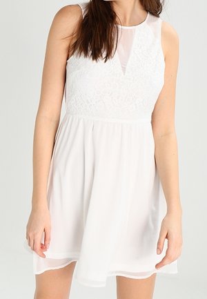 Biała sukienka bez rękawów z koronkową częścią górną i przezroczystą wstawką w kształcie litery V przy dekolcie, noszona przez osobę o długich brązowych włosach.