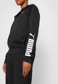 Sudadera con cremallera negra de tejido suave, con un logo blanco de Puma en el pecho y letras en negrita a lo largo de las mangas.