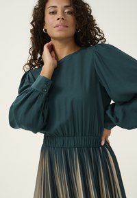 Green blouse with puff sleeves, elastic waist, and pleated beige skirt. Smooth texture, button details on cuffs, elegant silhouette.