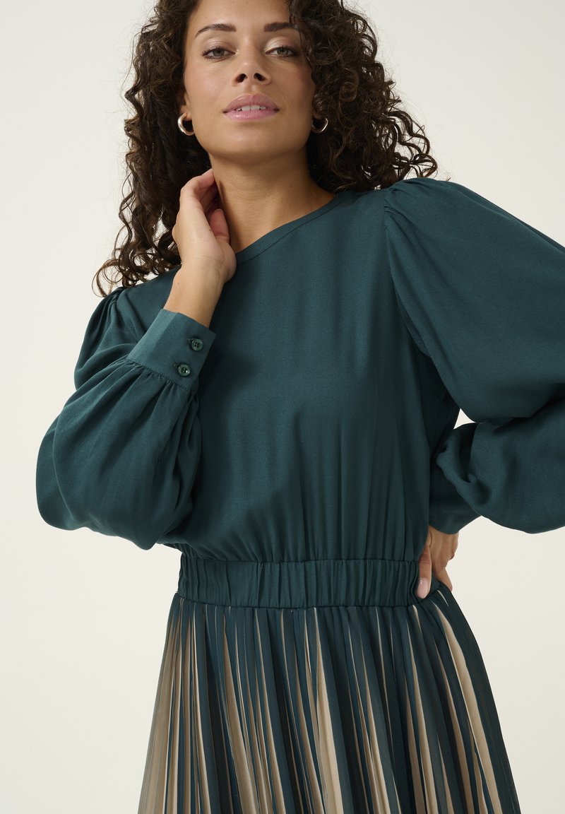 Blusa verde con mangas abullonadas, cintura elástica y falda beige plisada. Textura suave, detalles de botones en los puños, silueta elegante.