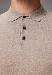 Beige pólóing texturált anyagból, gallér nélküli nyakkal, három fekete gombbal és letisztult, minimalista dizájnnal.