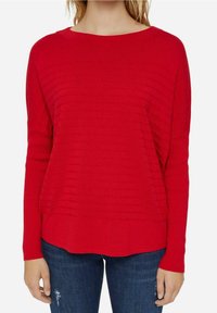 Kvinde iført en lys rød ribbestrikket sweater med vandrette strukturerede striber og mørkeblå slidte jeans mod en hvid baggrund.