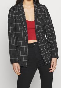 Black checked blazer with white grid pattern; features notch lapels, single button closure, and two front pockets. Worn over a red ribbed top.