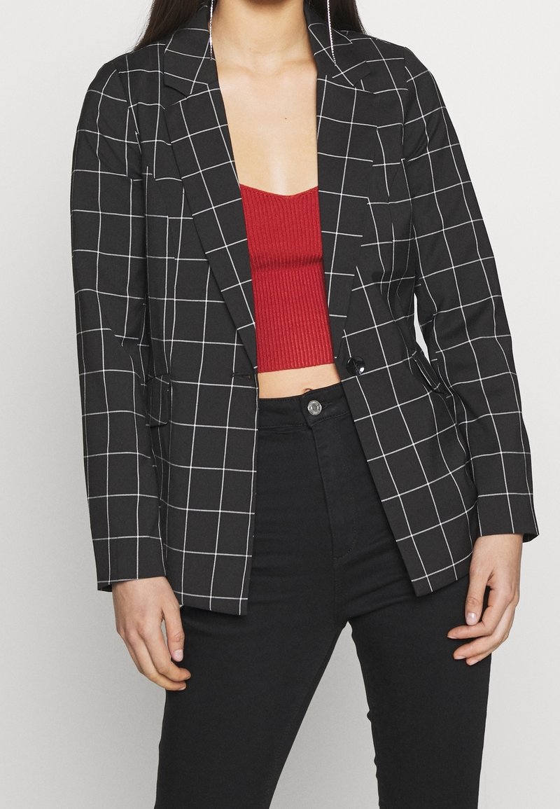 Black checked blazer with white grid pattern; features notch lapels, single button closure, and two front pockets. Worn over a red ribbed top.