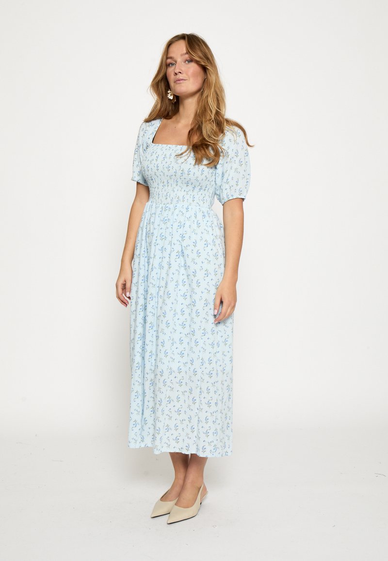 Mujer con el cabello largo que lleva un vestido midi azul claro con estampado floral, mangas abullonadas y escote cuadrado, zapatos puntiagudos beige, posando contra un fondo blanco.