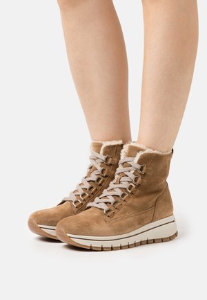 Brown suede ankle boots with faux fur lining, featuring beige laces and metal eyelets, and a thick white sole with textured grip.